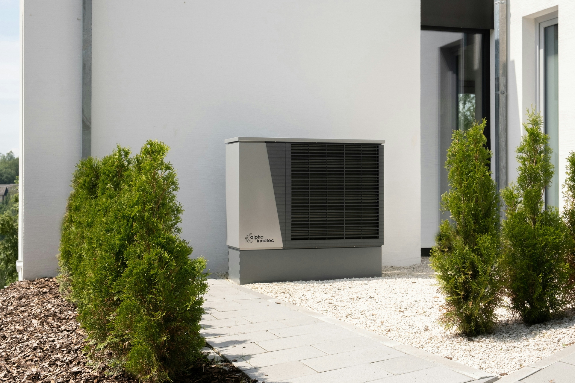 A modern outdoor heat pump unit installed beside a white building, surrounded by small green shrubs and a pathway made of square tiles.