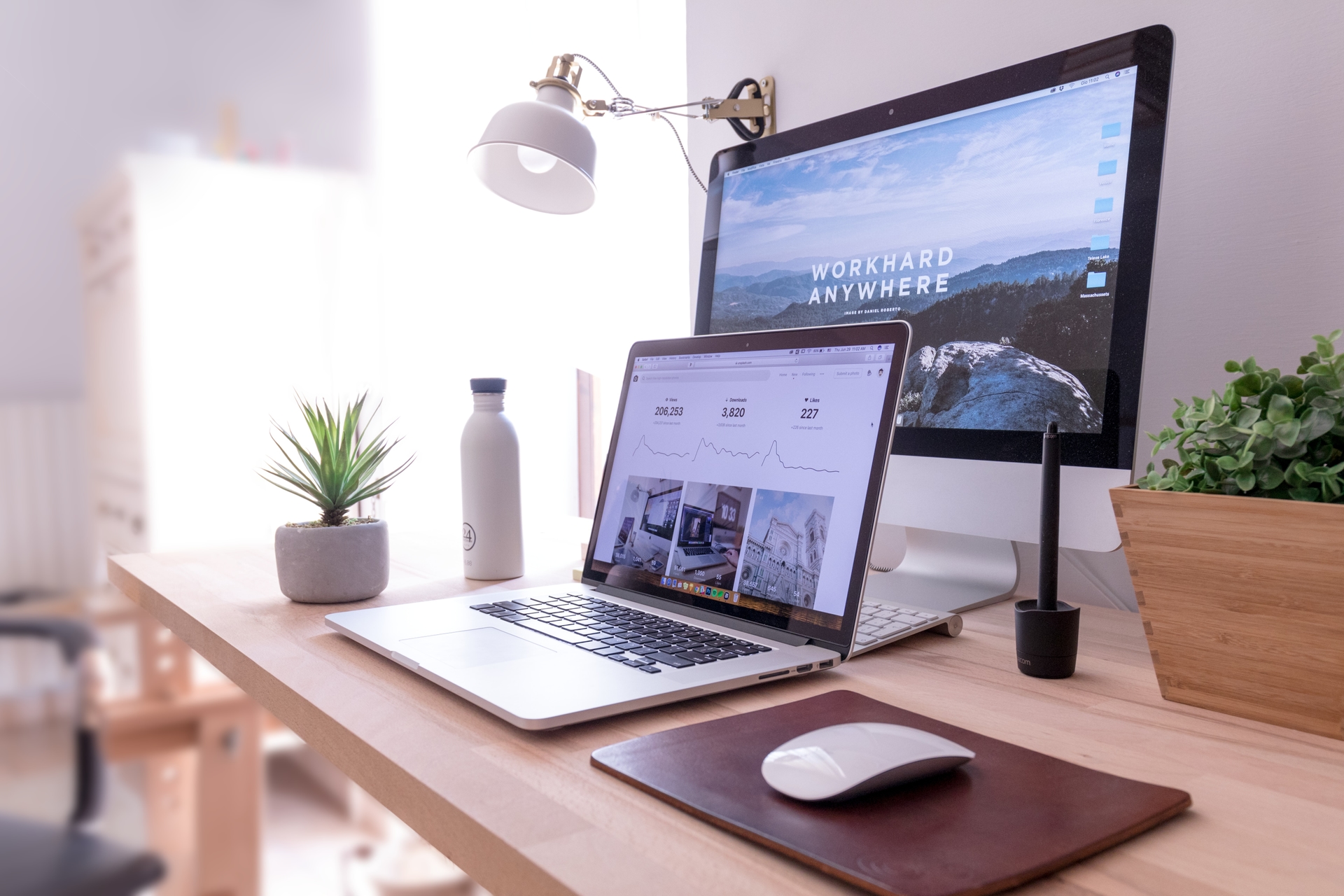 Minimalistischer Schreibtisch mit Laptop, großem Monitor, Zimmerpflanze, weißer Trinkflasche und Schreibtischlampe, auf dem Monitor der Schriftzug „Work Hard Anywhere“.